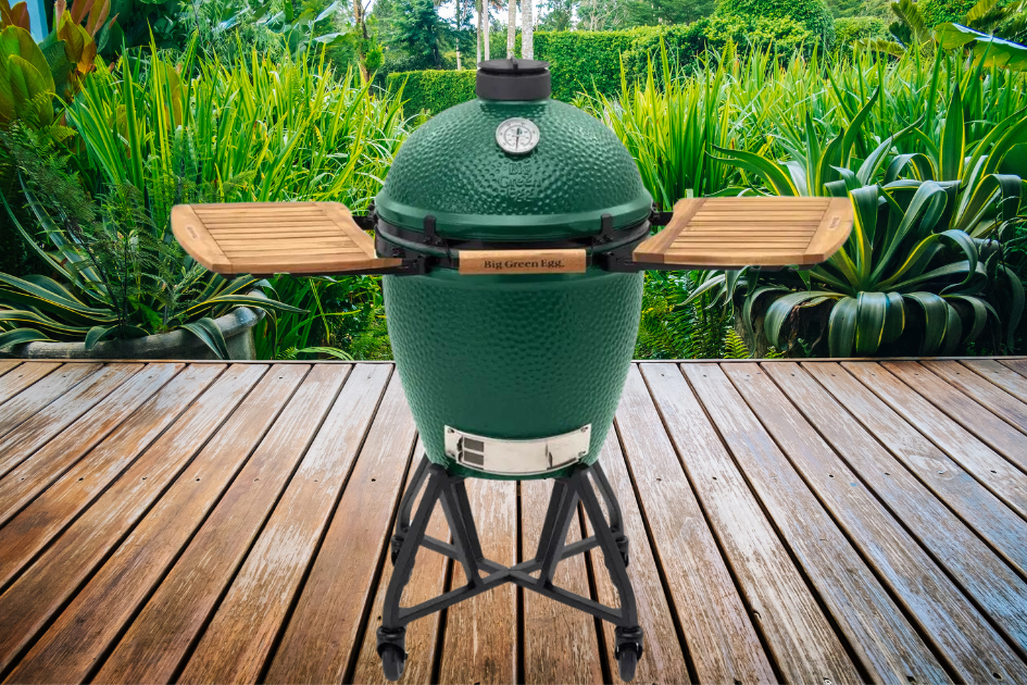 A Big Green Egg charcoal grill sits on a wood deck in a backyard