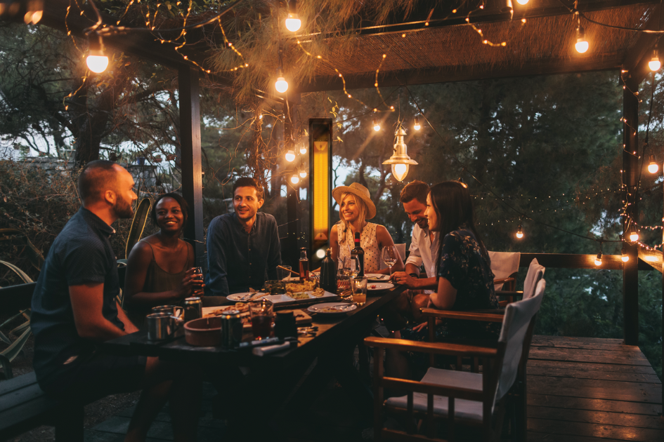 A group of friends sits around an outdoor table eating food and talking. String lights illuminate them from above. 