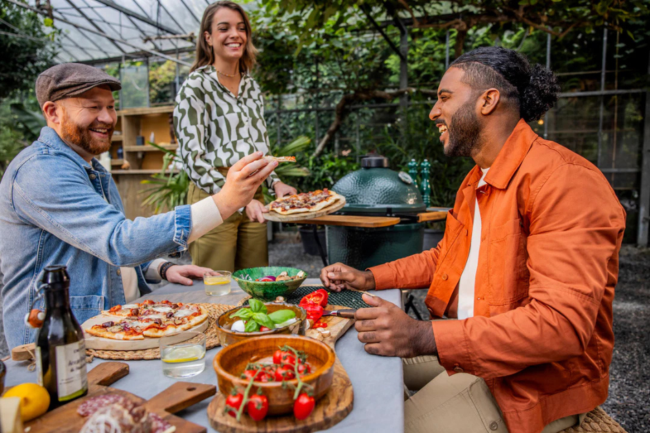 Three friends laugh and share BBQ around a Big Green Egg grill on their patio.