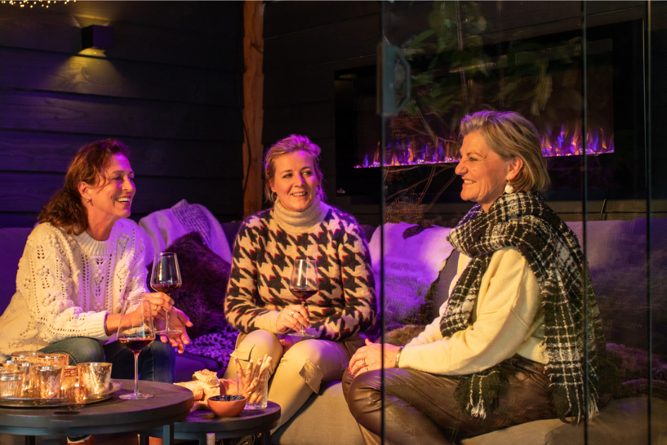 Three friends sit on a couch laughing together, with the warm glow of a Napoleon fireplace behind them 